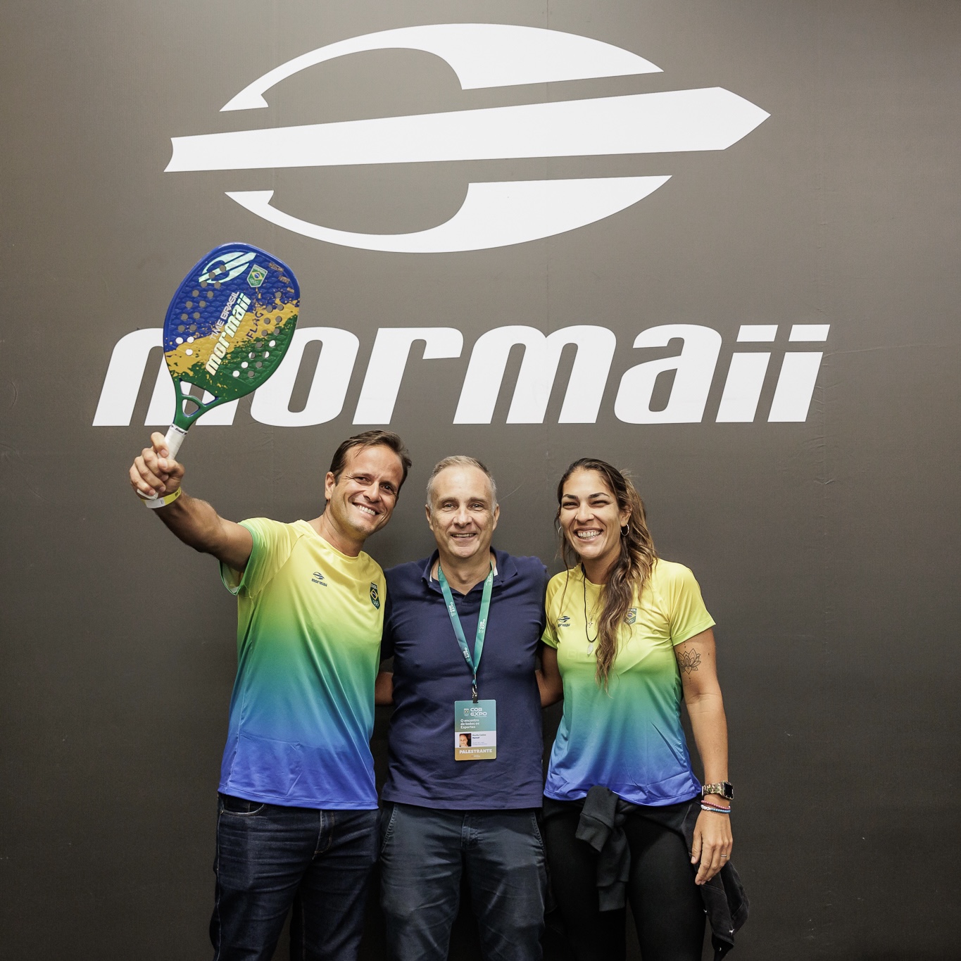 Atletas de Beach Tennis Mormaii com palestrante no evento da Cob Expo em São Paulo. Eles estão na frente de uma parede com a marca da Mormaii estampada.