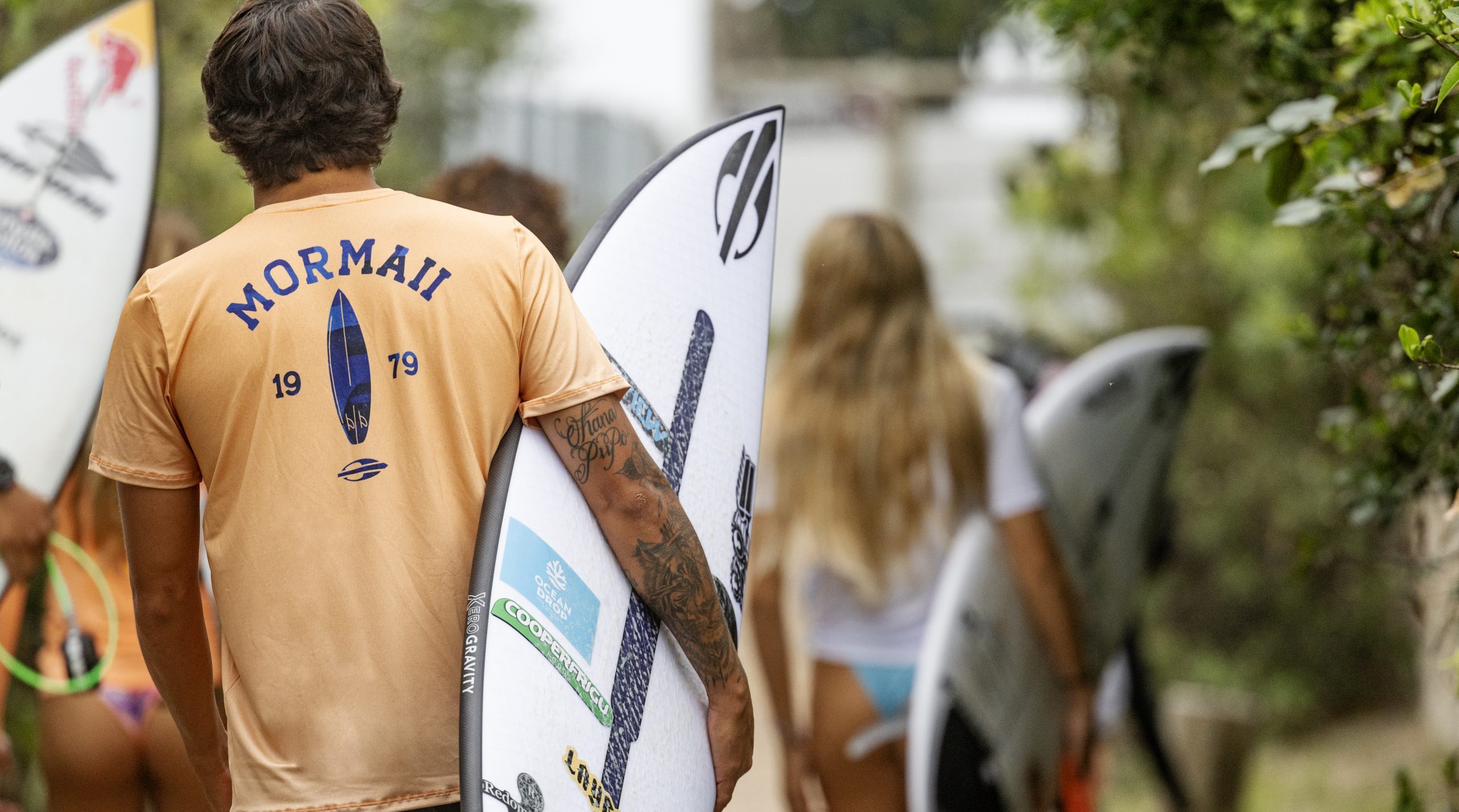 Surfistas de costas, carregando as pranchas e usando blusas com logo mormaii, fazendo alusão ao franchising